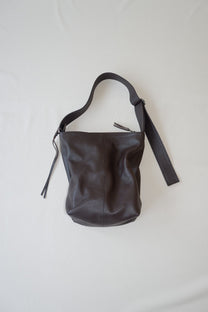 A dark brown leather shoulder bag with a slouchy, bucket-like silhouette, featuring a matching leather strap and small tassel details, laid flat on a light off-white fabric background.